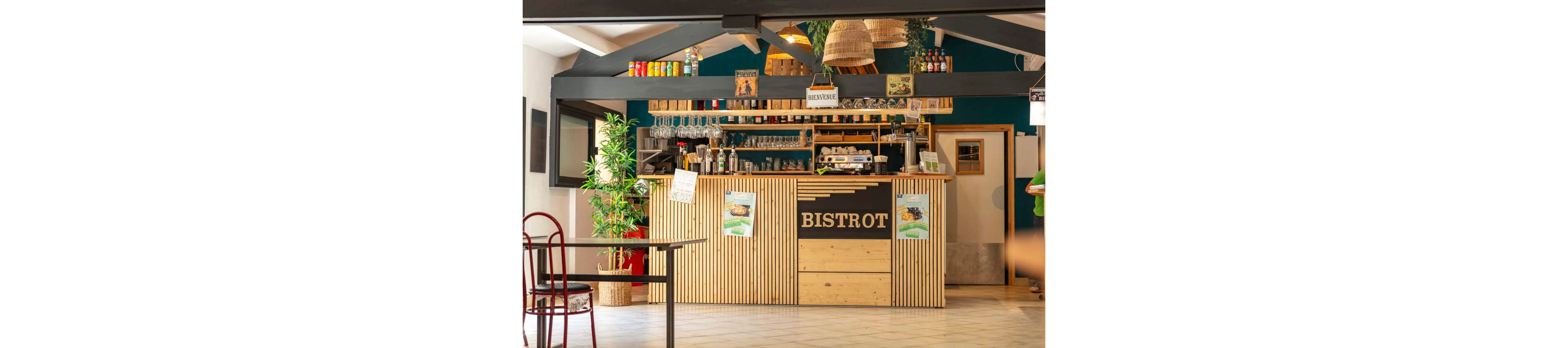 A warm, inviting bistro bar features a wooden slat counter with the word "BISTROT" prominently displayed on the front. Shelves behind the bar hold bottles, glasses, and a coffee machine, creating a casual café atmosphere. Hanging wicker lamps and green plants add natural texture and a relaxed ambiance. A small sign reading "BIENVENUE" welcomes guests into the bright, open space.
