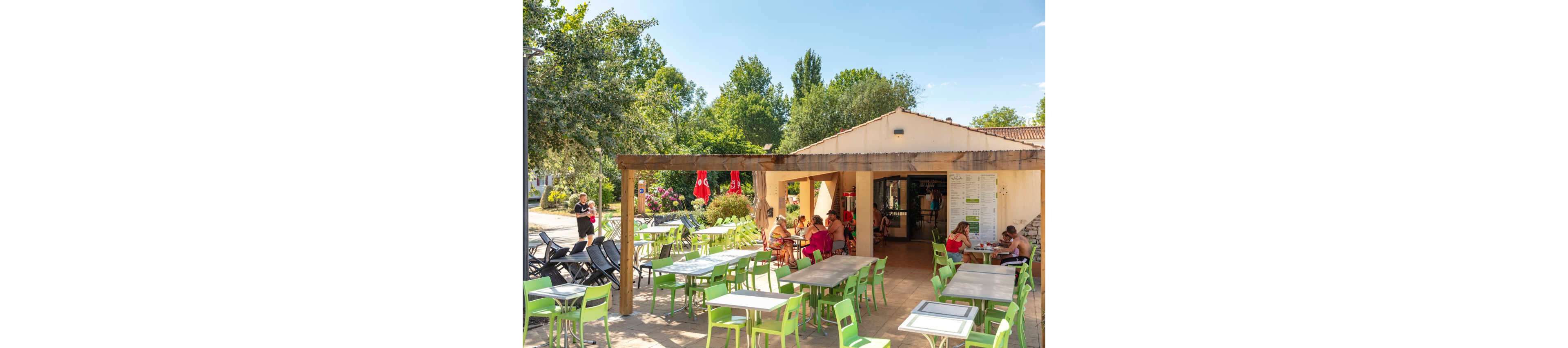 An open-air café terrace with green and white tables and chairs is set beside a small building in a sunny outdoor setting. People sit and chat under the shaded pergola while others walk nearby, creating a relaxed holiday atmosphere. Lush green trees and flowering bushes surround the café, suggesting a campsite or vacation resort. Bright red parasols and clear blue sky emphasize the warm summer weather.