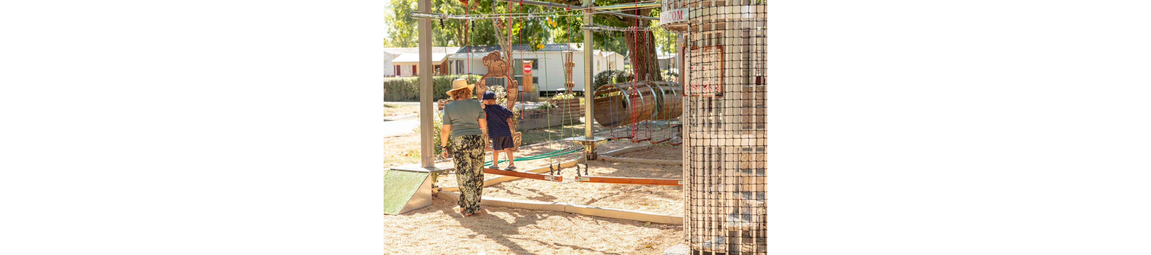 An older adult walks beside a young child on a rope and balance playground structure. The scene takes place outdoors on a sunny day in a park or campground with mobile homes in the background. The child is holding onto ropes while navigating the play equipment, and the adult offers support from behind. Warm sunlight, trees, and sandy ground create a relaxed, family-friendly atmosphere.