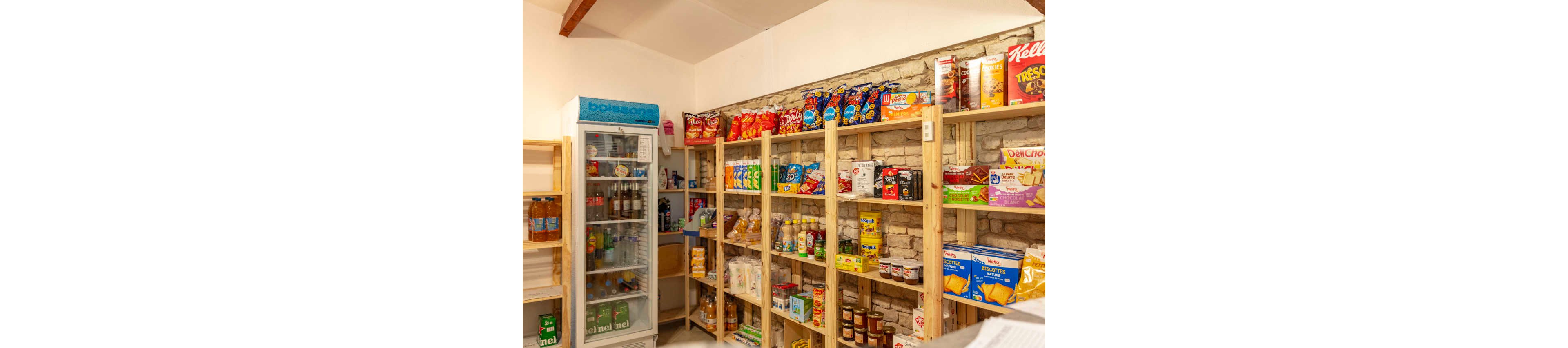 Interior of a small grocery or convenience store with wooden shelves filled with assorted snacks and dry goods. Colorful bags of chips and boxes of cereal line the upper shelves, while canned foods, pasta, and other pantry items occupy the lower levels. On the left side, a glass-door refrigerator displays bottled drinks and beverages. The setting has a warm, rustic feel with exposed brick behind the shelving and simple overhead lighting.