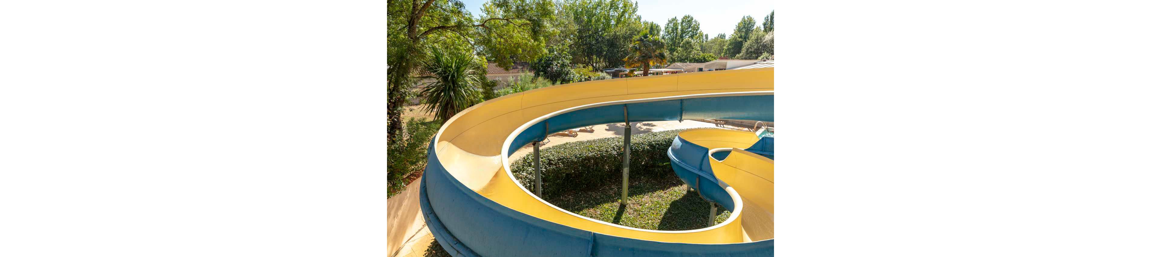 A young child rides down a large curved waterslide at an outdoor water park. The slide is yellow on the inside with blue outer sections, winding above grass and shrubs. Sunlight filters through surrounding green trees, creating a bright summer atmosphere. The scene feels playful and energetic, capturing a fun vacation or holiday moment.