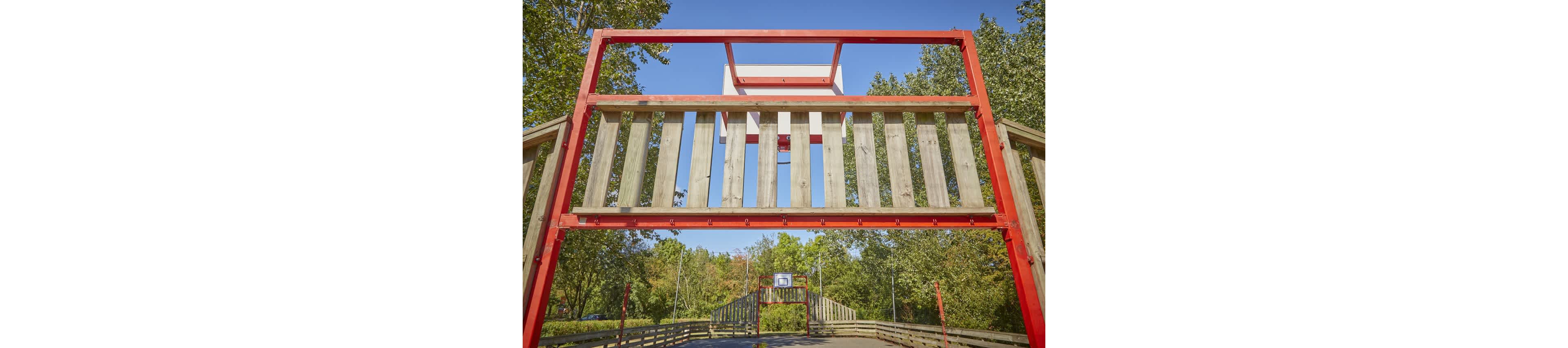 An outdoor basketball court features multiple wooden and red metal structures with hoops aligned in a row. The image is taken from beneath a large wooden frame, looking down the length of the court. Trees with green foliage surround the court, suggesting a park or recreational area. The bright daylight and clear sky create a warm, inviting atmosphere for outdoor sports.
