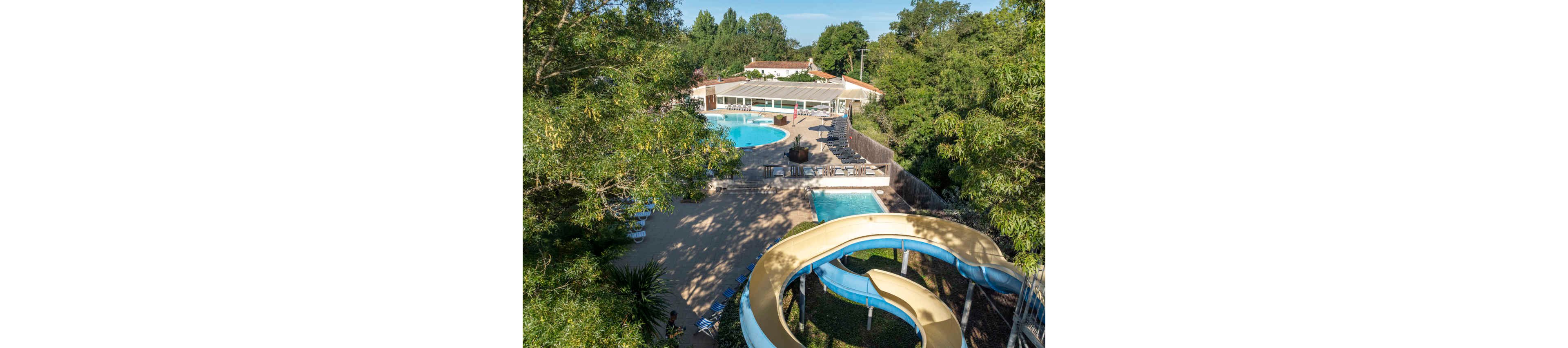 A large outdoor swimming pool complex is surrounded by trees and greenery. The scene includes a curving blue and white waterslide leading to a small pool. Sun loungers and umbrellas are arranged around the main pool near a low building with a glass-fronted area. The setting appears to be a leisure resort or campground on a sunny day, with clear blue water and bright light creating a relaxed vacation atmosphere.