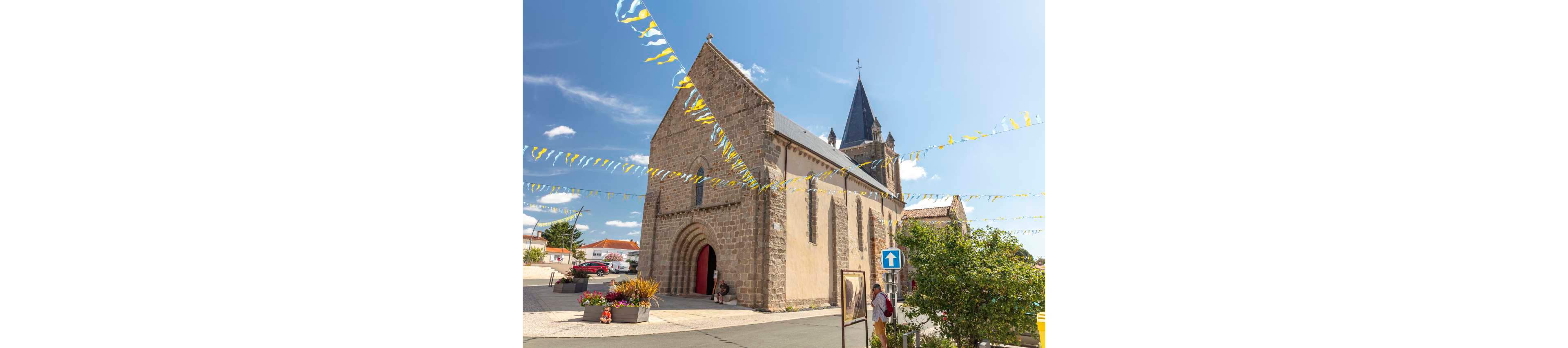 A historic stone church stands on a sunny village street, its arched entrance and tall bell tower dominating the scene. Colorful triangular bunting is strung across the sky, adding a festive touch to the quiet square. A few people walk or sit near the church, while potted flowers and shrubs brighten the foreground. A blue one-way traffic sign and surrounding houses indicate a small European town setting under a clear blue sky.