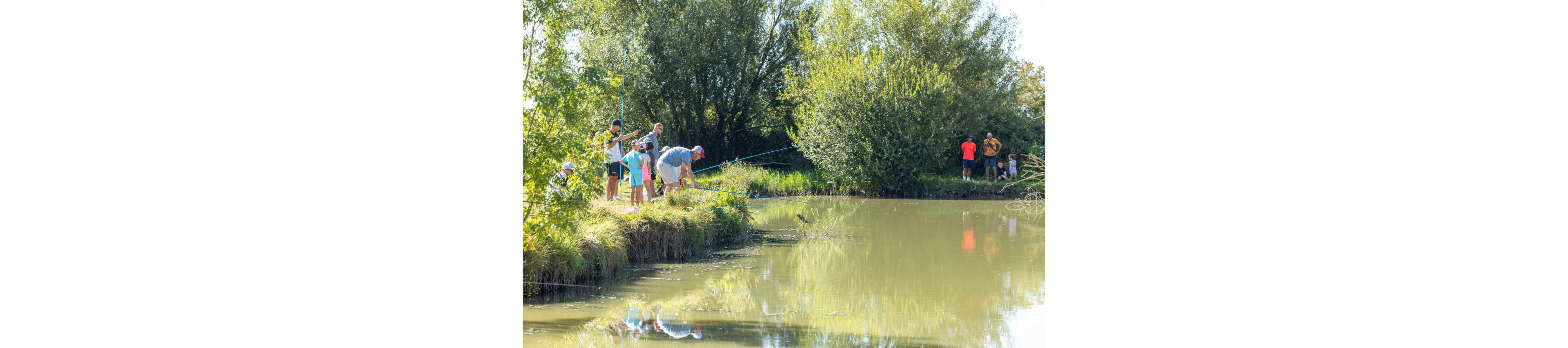 A group of people gather along the grassy edge of a calm lake to fish on a sunny day. Adults and children stand close to the water, some holding fishing rods while others watch the activity. More people are visible in the background near the trees, enjoying the outdoor setting. Lush green foliage and reflections on the water create a peaceful, relaxed atmosphere.