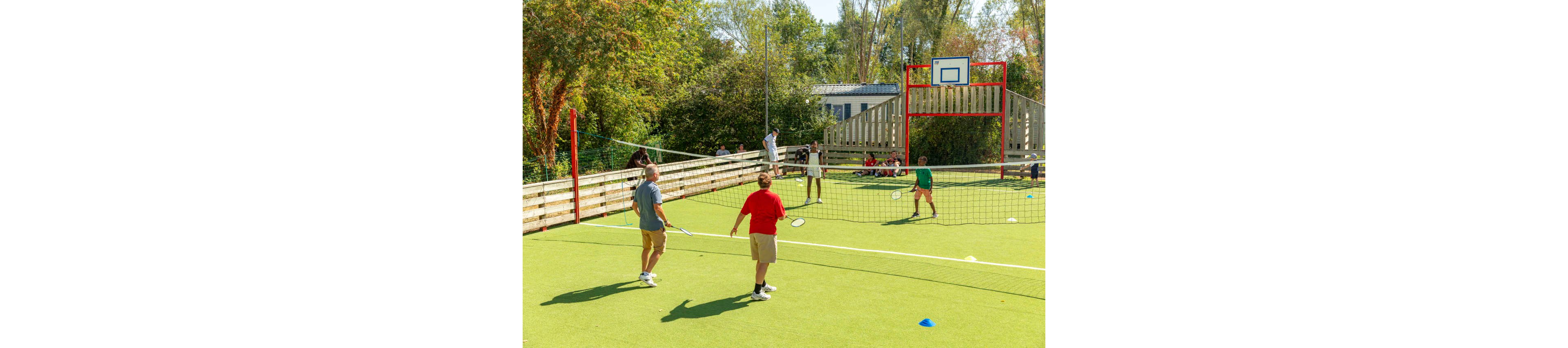 Several people play badminton on a bright green outdoor court. A net stretches across the middle while players stand on both sides holding rackets. In the background, more people sit and watch near a wooden fence and a small building. A basketball hoop stands above the fence, and colorful cones mark areas on the court. Trees and greenery surround the recreational sports area under clear daylight.