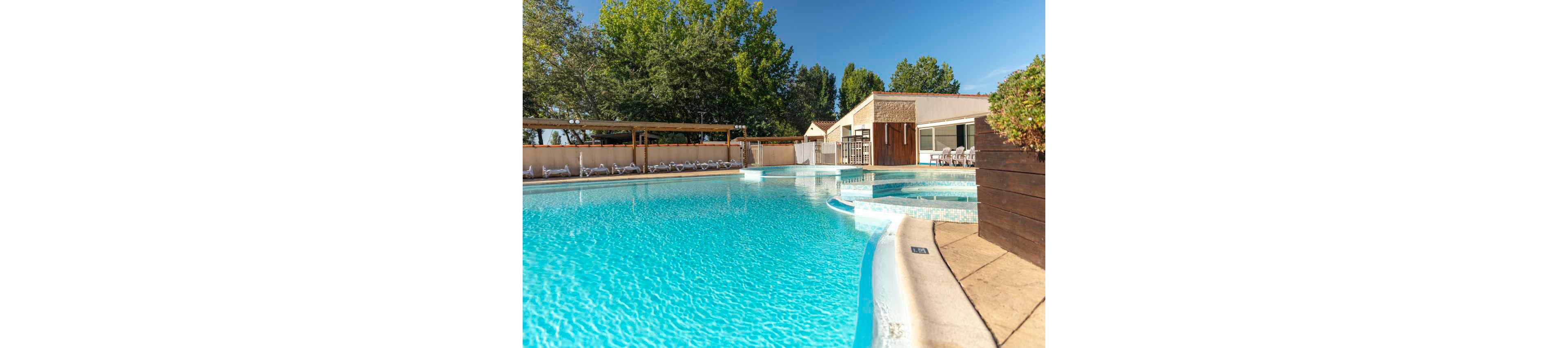 A large outdoor swimming pool glistens under clear blue skies at a resort or vacation property. The pool features curved edges and an attached circular spa area with steps leading into the water. Sun loungers are lined up along the far side beneath a shaded pergola structure. Modern low-rise buildings and lush green trees frame the relaxing leisure setting. The bright sunlight and turquoise water create a warm, inviting holiday atmosphere.