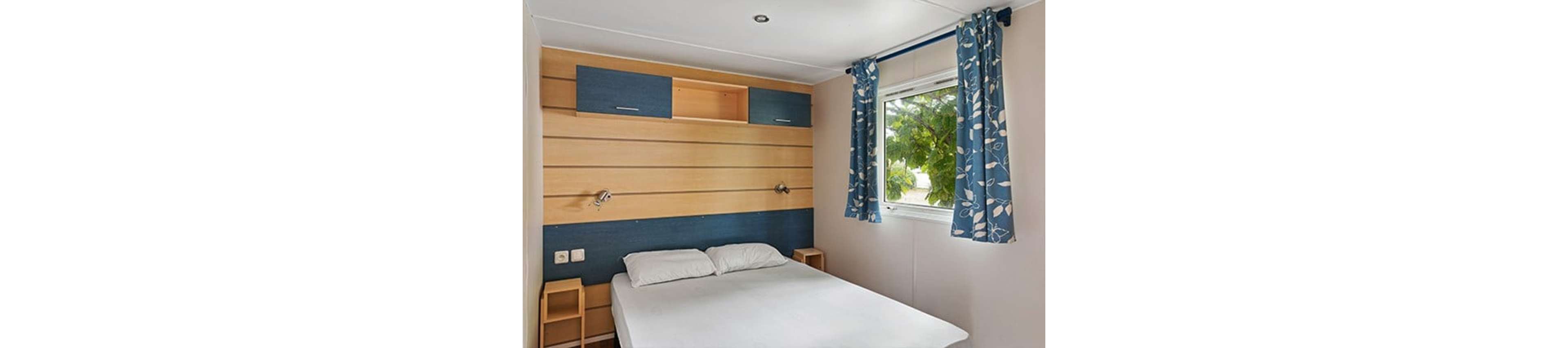 A small bedroom features a neatly made double bed with white linens against a light wooden accent wall. Built-in blue and wood storage cabinets hang above the bed, with small bedside shelves on each side. A window with blue patterned curtains lets in natural light and offers a view of green foliage outside. The overall design is simple, modern, and functional, creating a cozy and inviting sleeping space.