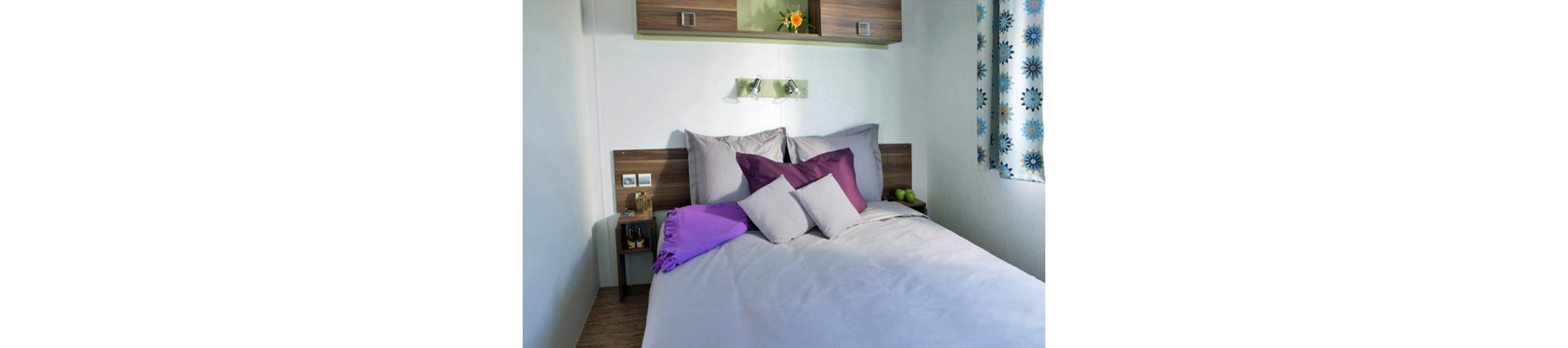 A neatly made bed with gray linens and purple and gray accent pillows is the main focus of this modern bedroom. Wooden headboard panels and small bedside shelves frame the bed, holding decorative items and a couple of green apples. Above the bed, a small shelf with a flower arrangement and wall-mounted reading lights adds visual interest. A patterned curtain with blue and yellow flowers brings additional color to the otherwise neutral room.