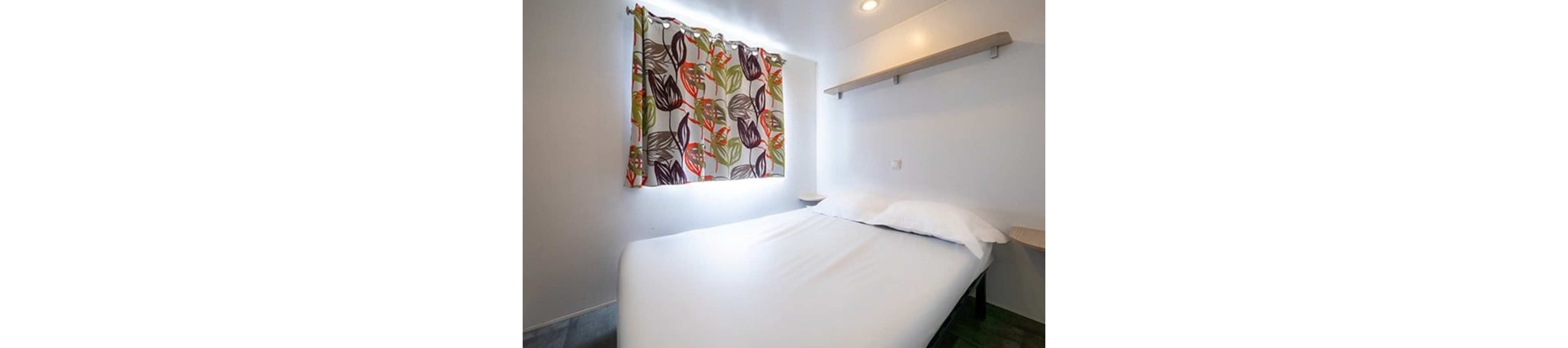 A compact bedroom features a neatly made double bed with white linens against a plain white wall. A small window is covered by colorful leaf-patterned curtains that add a decorative accent to the simple space. A narrow floating shelf is mounted above the bed, providing minimal storage or display area. Recessed ceiling lighting brightens the clean, modern room.