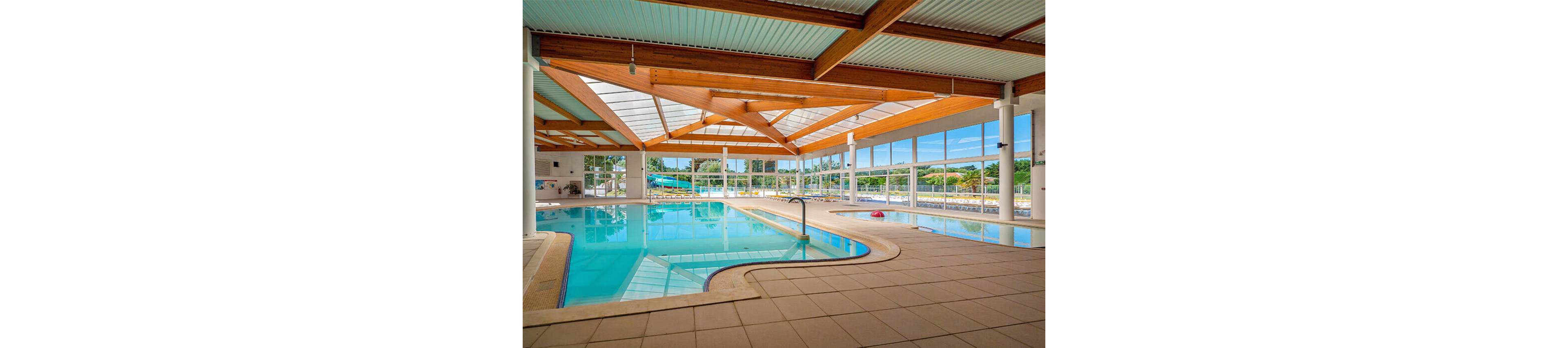 Spacious indoor swimming pool with clear blue water sits beneath a modern wooden beam ceiling and large skylights. Floor-to-ceiling windows surround the pool area, revealing green trees and an outdoor slide in the background. The tiled pool deck curves around the water with stainless steel handrails and a few scattered pool floats. Natural daylight fills the space, creating a clean, inviting atmosphere for recreation and relaxation.