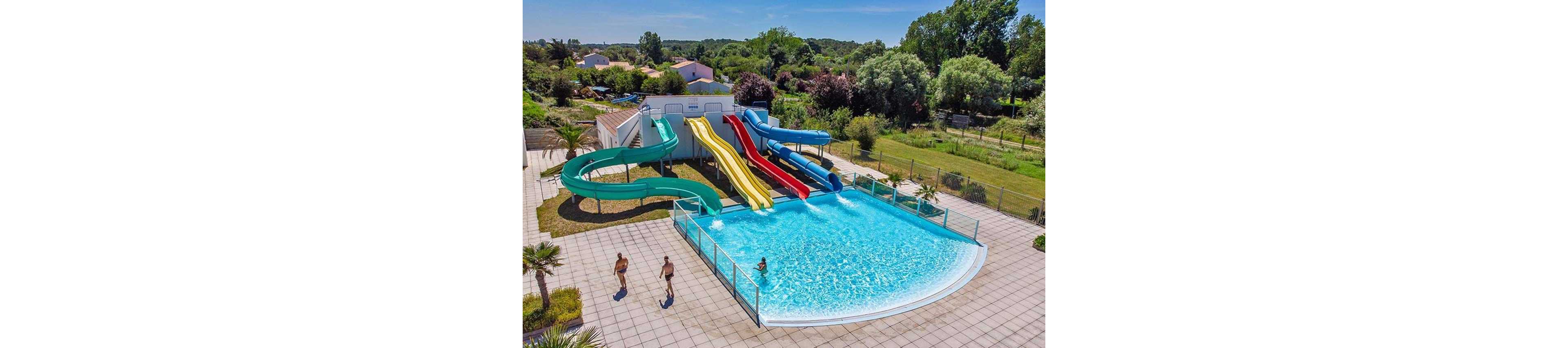 An outdoor water park features a large clear blue swimming pool with multiple colorful water slides emptying into it. The slides include green, yellow, red, and blue chutes curving and dropping from a raised platform. Two people walk along the tiled pool deck, while lush green trees and grass surround the fenced area. Nearby buildings and a distant tree line indicate a recreational complex in a suburban or rural setting.