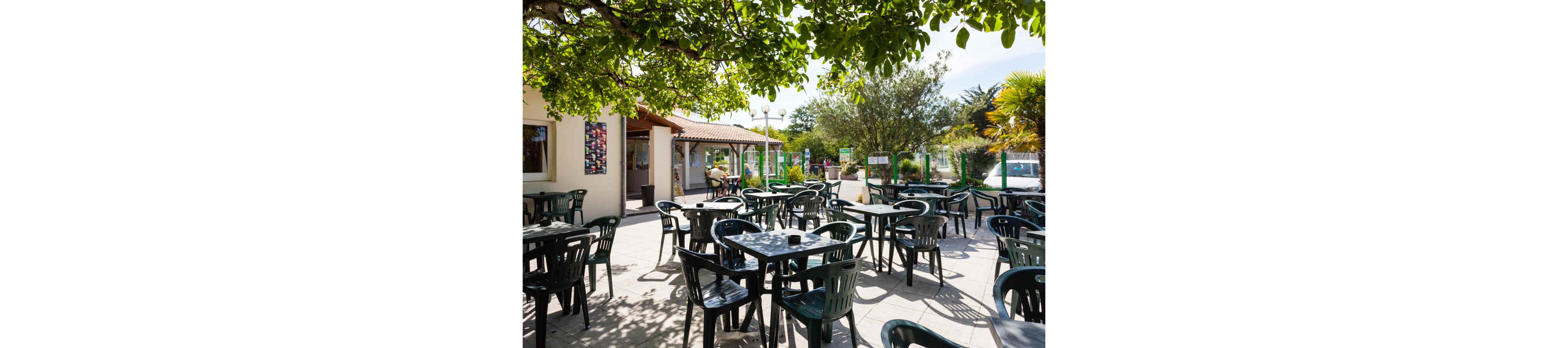 An outdoor café terrace is filled with empty black plastic tables and chairs under the shade of leafy trees. Sunlight creates bright patches on the tiled patio, suggesting a warm, pleasant day. A low building with large windows and colorful posters forms the backdrop, with additional seating and greenery extending into the distance. The scene feels calm and inviting, ready for guests to arrive.