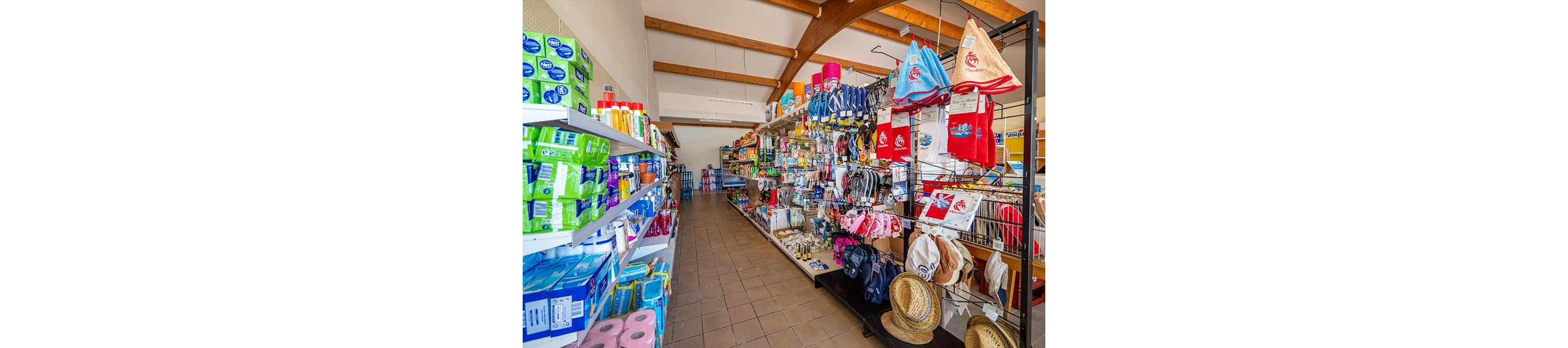 A bright aisle in a small convenience store is filled with various household and personal care products. Shelves on the left display toiletries, cleaning supplies, and paper goods, while racks on the right hold hats, bags, towels, and beach accessories. The tiled floor and wooden ceiling beams lead the eye toward the back of the shop, where more products are stacked. The overall scene suggests a well-stocked local market catering to everyday needs and vacation shoppers.