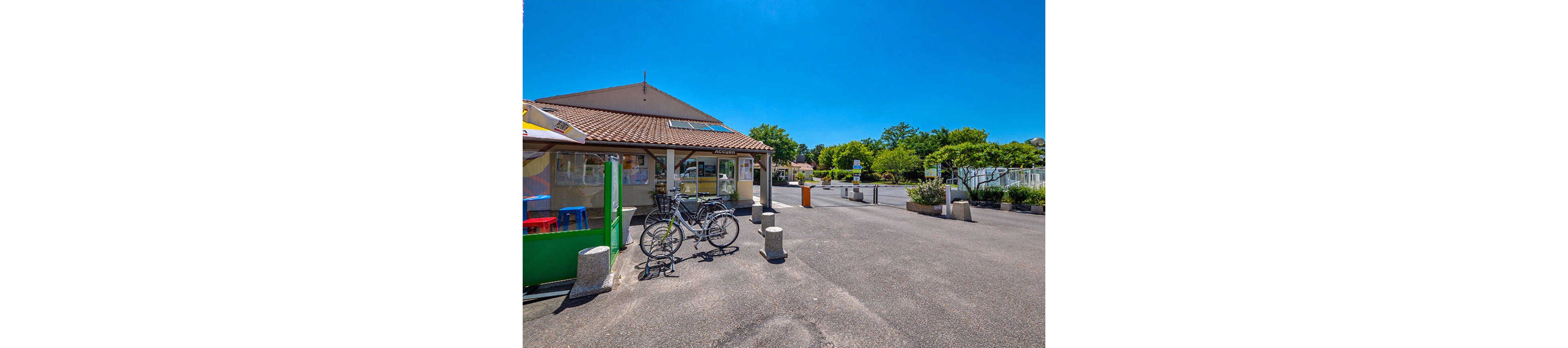 A small single-story building with a tiled roof stands beside an open paved courtyard under a bright blue sky. Two bicycles are parked near concrete bollards in the foreground, suggesting a casual stop or visit. Colorful outdoor chairs and a kiosk-like structure appear on the left, adding a touch of vibrancy. Trees and additional structures are visible in the background, giving the scene a relaxed, suburban or holiday park atmosphere.