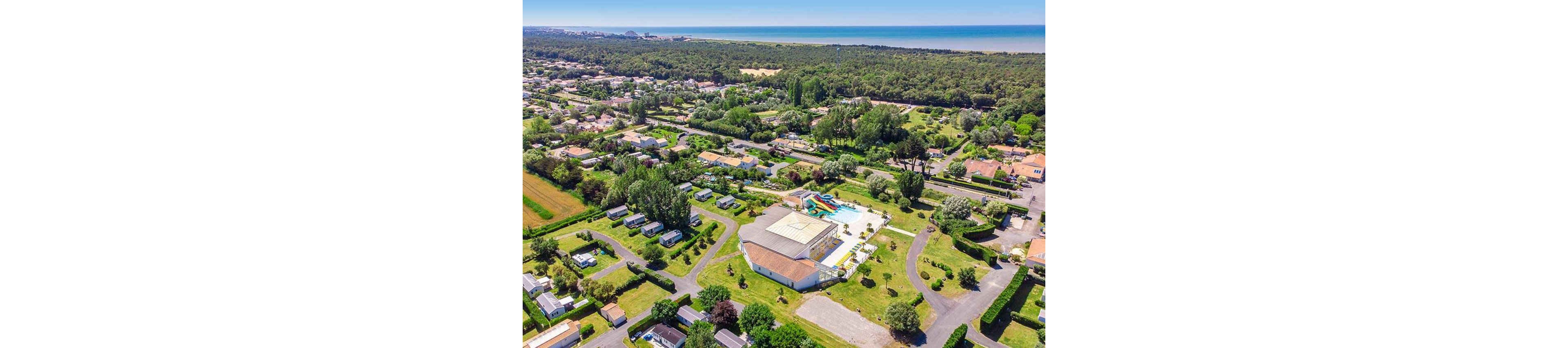 A large holiday campground with mobile homes and cabins is seen from above near a forested coastline. The central complex features an outdoor swimming pool area with colorful water slides and sun loungers. Curving internal roads connect neatly arranged plots of accommodation surrounded by green lawns and trees. In the distance, dense woodland stretches toward a sandy shoreline and calm sea under a clear sky.