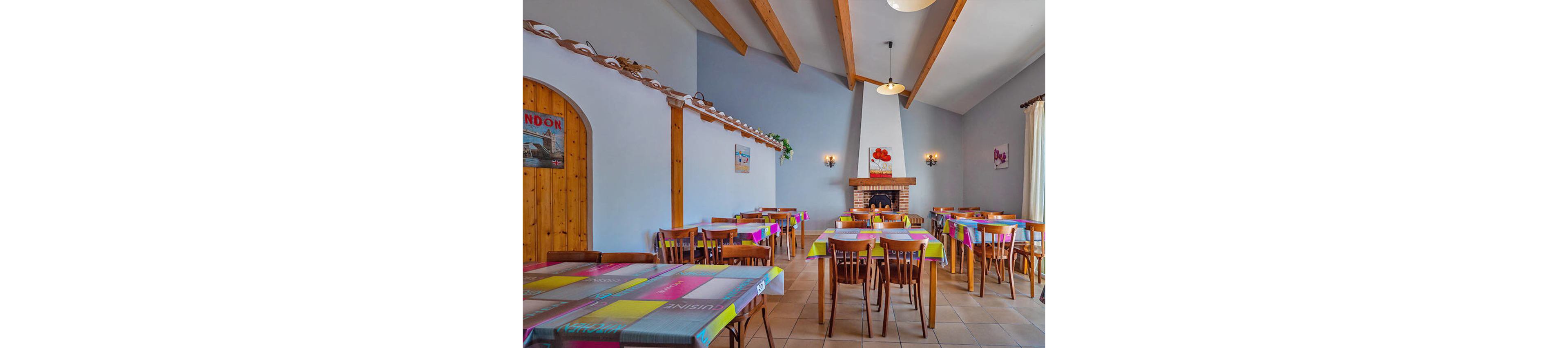 Brightly lit dining room with multiple wooden tables and chairs is arranged neatly around a central fireplace. Colorful tablecloths in bold geometric patterns add vibrancy to the neutral walls and tiled floor. Wooden ceiling beams and a brick fireplace create a cozy, rustic atmosphere. Wall art, sconces, and a large window with curtains complete the inviting restaurant-style interior.