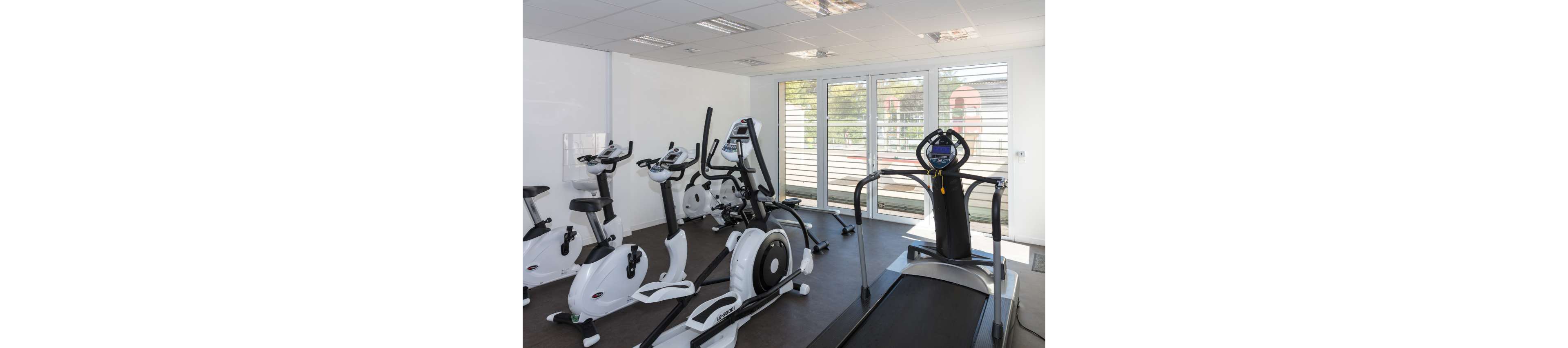 A clean, modern gym room features several white stationary bikes and an elliptical trainer arranged along one side. A treadmill with a digital console stands in the foreground facing large windows. Natural light floods the space through floor-to-ceiling glass doors with blinds, creating an airy and inviting workout environment. The room has white walls, a suspended ceiling with lights, and dark flooring that contrasts with the fitness machines.