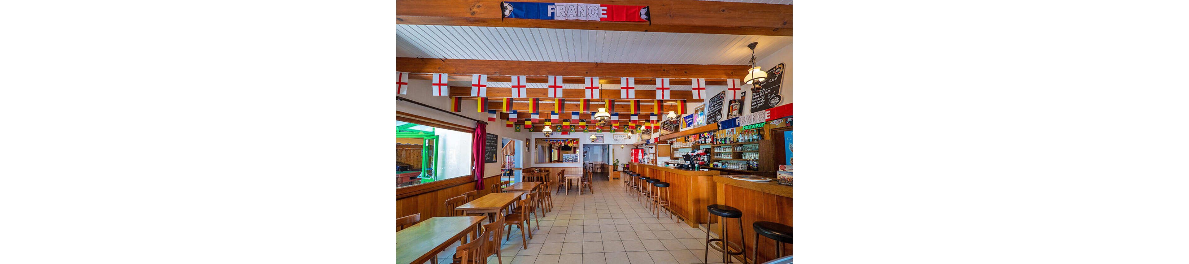 A bright, spacious bar interior is decorated with numerous hanging national flags, including many with red crosses on white backgrounds. Wooden tables and chairs line the tiled floor, while a long wooden counter with bar stools runs along the right side. Chalkboard menus and shelves of bottles are visible behind the bar, and a banner reading "FRANCE" hangs from the ceiling beam. The overall atmosphere suggests a casual venue prepared for watching international sports events.