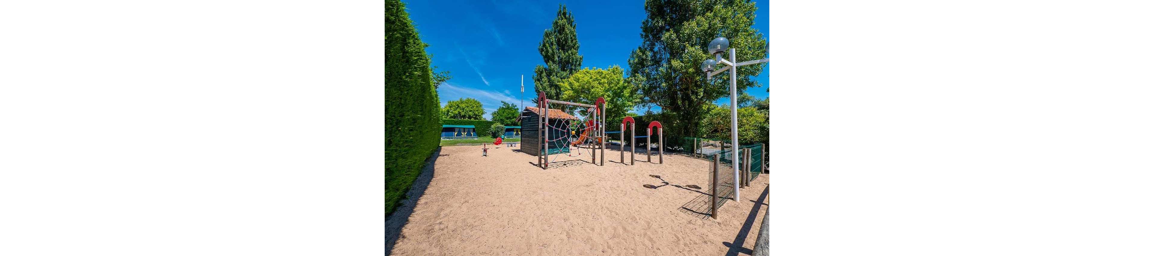 An empty sandy playground features a central climbing structure with a rope web and curved red bars. The play area is surrounded by green hedges, trees, and a wire fence under a bright blue sky. In the background, additional play equipment and shaded seating can be seen. The scene suggests a quiet, sunny day in a park or campground recreation area.