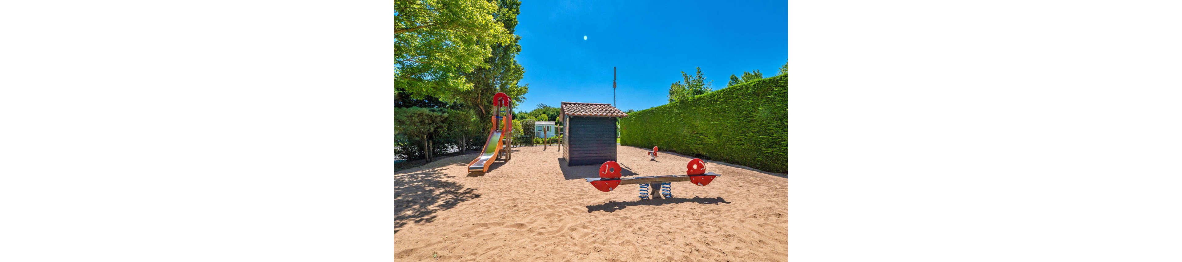 An empty sandy playground is shown under a bright blue sky. A colorful slide and climbing structure stand to the left, while a small playhouse sits in the center. In the foreground, a red and blue spring seesaw is positioned on the sand. Green hedges and trees surround the play area, creating a private and inviting outdoor space.