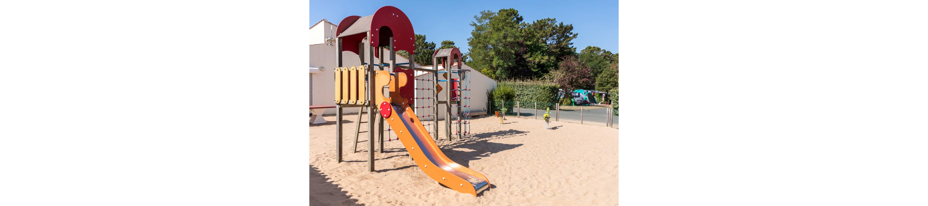 An empty outdoor playground features a colorful slide and climbing structure set in a large sand area. The play equipment includes a red and orange slide, ladders, and rope climbing nets. The playground is surrounded by a low fence, with trees, shrubs, and nearby buildings in the background. Bright daylight and clear blue sky create a warm, inviting atmosphere.