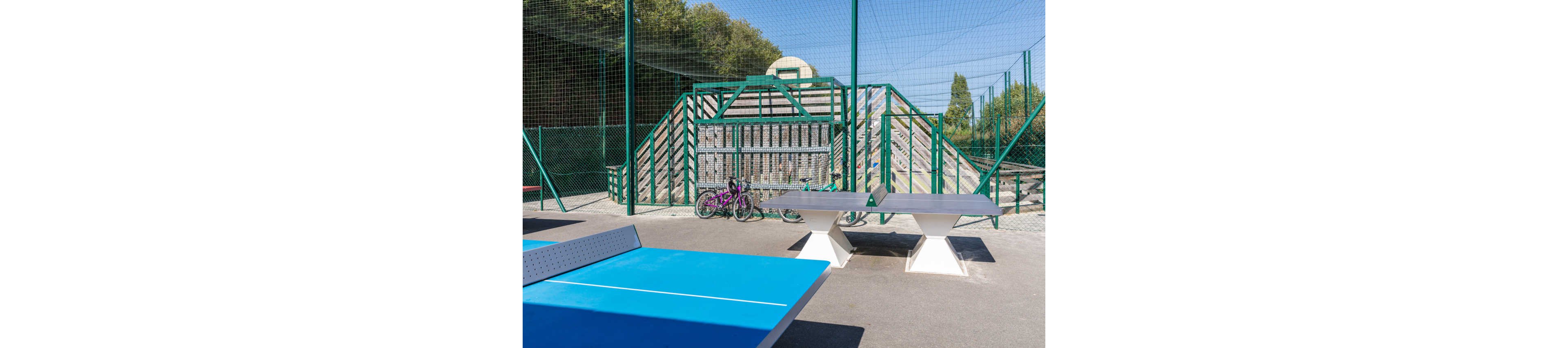 An outdoor recreational sports area features two blue table tennis tables in the foreground. Behind them, several bicycles are parked against a green metal fence and a wooden climbing structure. The space is enclosed by high netting and fencing, suggesting a safe play zone. Trees and greenery surround the court, indicating a park or community sports facility on a sunny day.