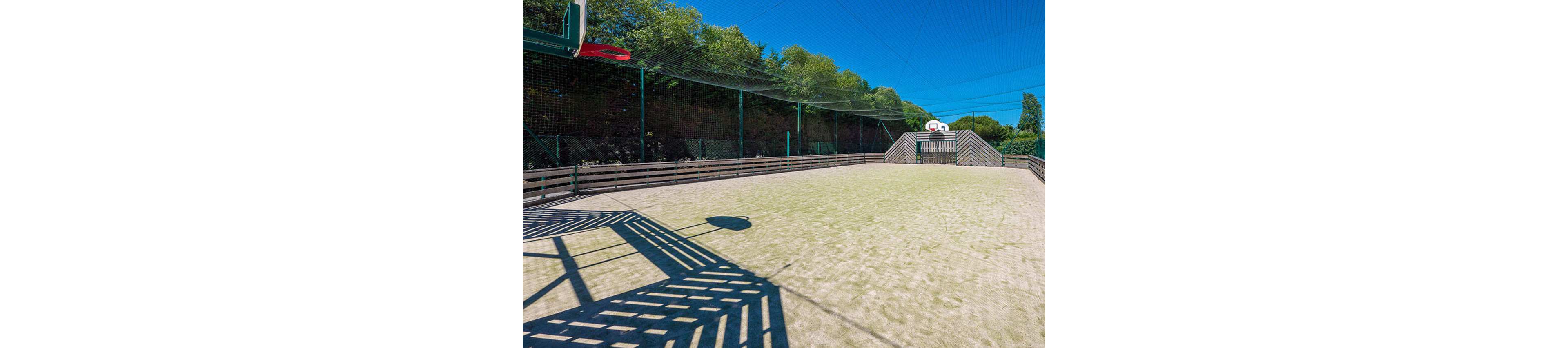 An empty outdoor multi-sport court is shown under a clear blue sky. The fenced area features basketball hoops at each end and wooden barriers along the sides. Netting stretches overhead, and strong sunlight casts geometric shadows on the light-colored playing surface. Trees and greenery form a natural backdrop behind the court, creating a bright and open recreational setting.