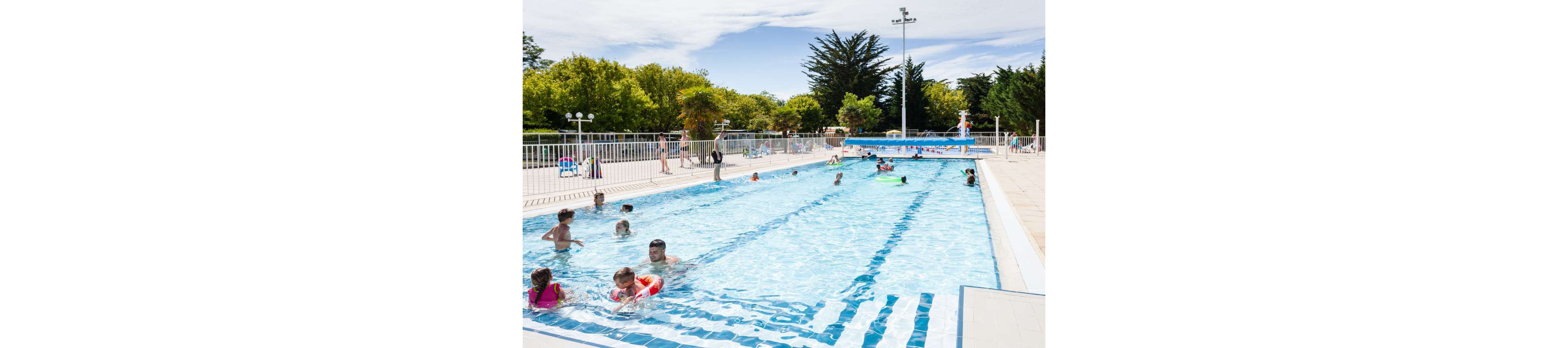An outdoor swimming pool is filled with people enjoying a sunny day. Children and adults play and swim in the clear blue water near the pool edge and in the deeper section. White safety railings and poolside deck chairs surround the pool area. Lush green trees and a bright sky create a relaxed, summery atmosphere.