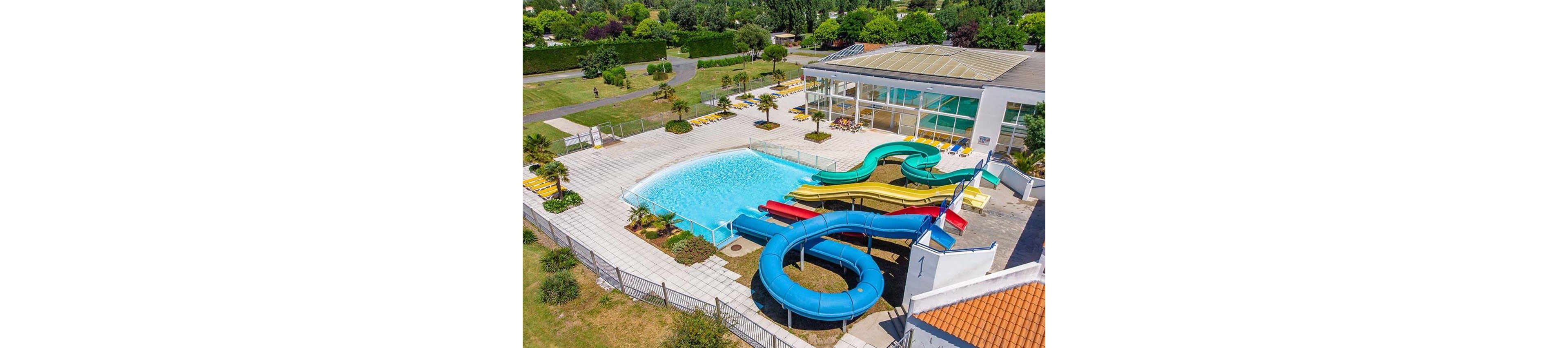 A colorful outdoor water park features a large swimming pool with multiple winding water slides. The scene is captured from above, showing the pool area surrounded by sun loungers and a fenced terrace. A modern building with large glass windows and an indoor pool area stands beside the main pool. Lush green lawns, trees, and walking paths extend into the background, suggesting a spacious recreational complex.