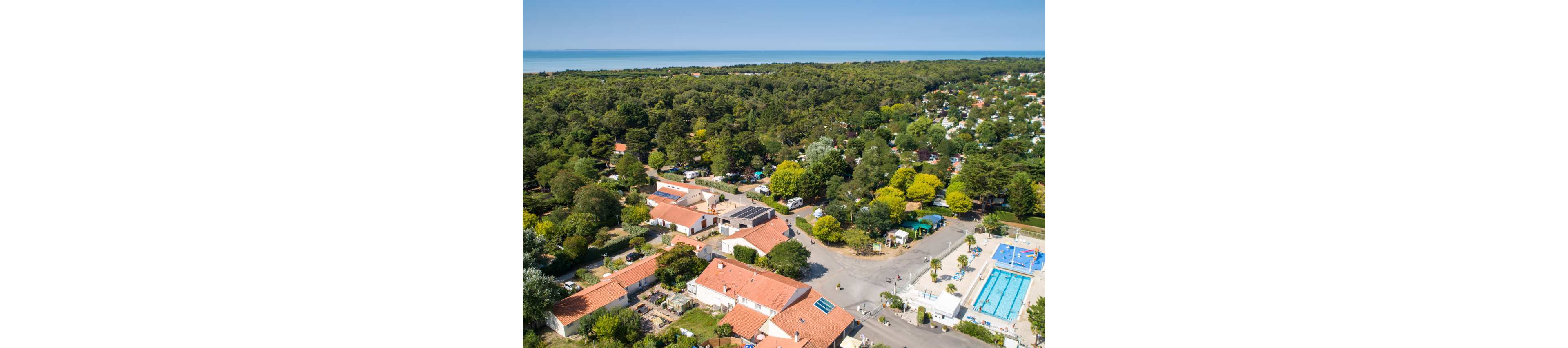 Aerial view of a coastal holiday campsite surrounded by dense green forest. The scene shows red-roofed buildings, caravans, and tents spread across the property. A large rectangular outdoor swimming pool and adjacent facilities are visible near the entrance area. In the distance, the blue sea meets the horizon, emphasizing the campsite’s seaside location.