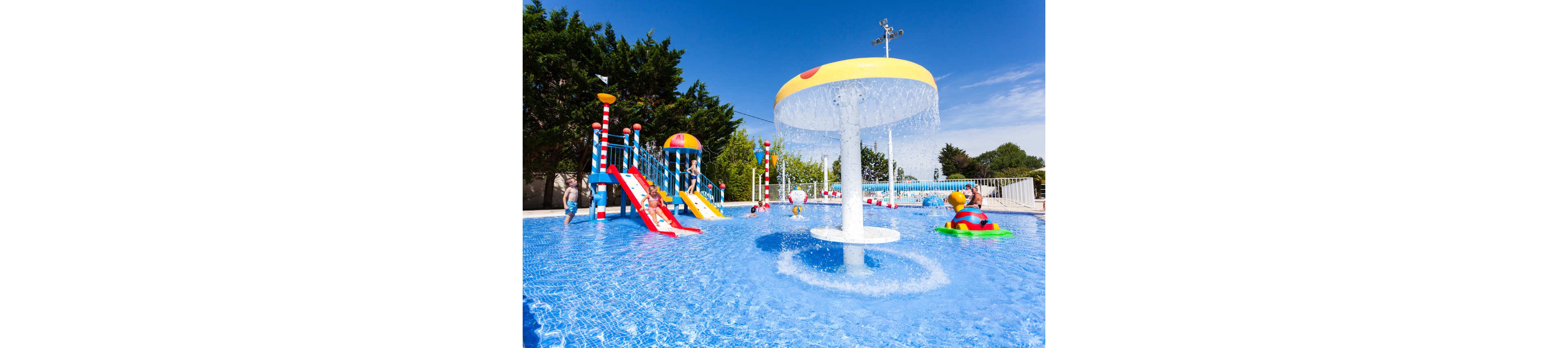 Children play in a shallow outdoor splash park filled with bright blue water. A large mushroom-shaped fountain pours water into the pool while kids climb and slide on colorful play structures. Small slides, ladders, and water toys create an energetic and fun atmosphere. The scene is set under a clear blue sky, suggesting a warm, sunny day at a family-friendly water park.