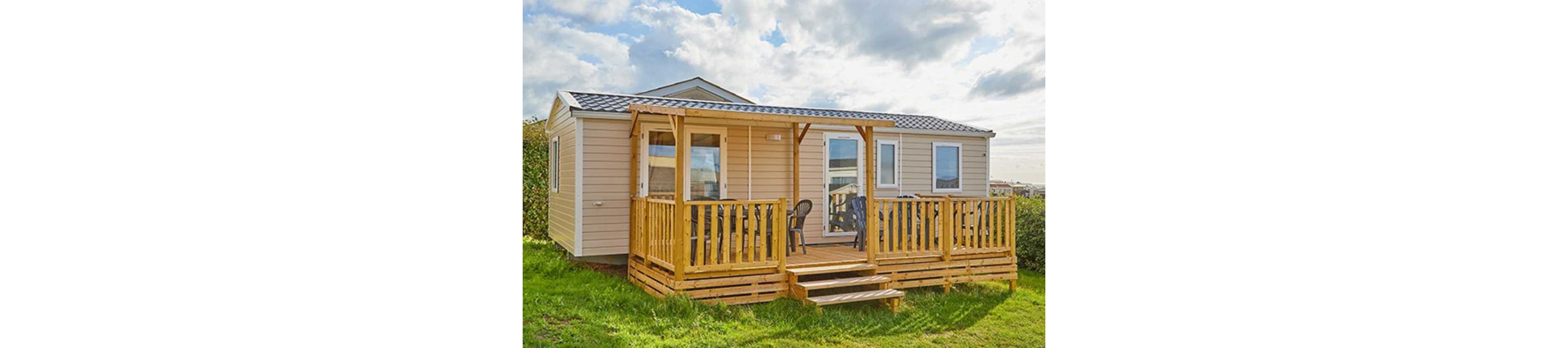 A beige mobile home with a pitched roof stands on a grassy plot under a partly cloudy sky. The front features a wooden deck with railings and steps leading up to the entrance. Plastic chairs are arranged on the porch, suggesting a relaxed outdoor seating area. In the background, other similar structures and greenery indicate a holiday park or residential campsite setting.