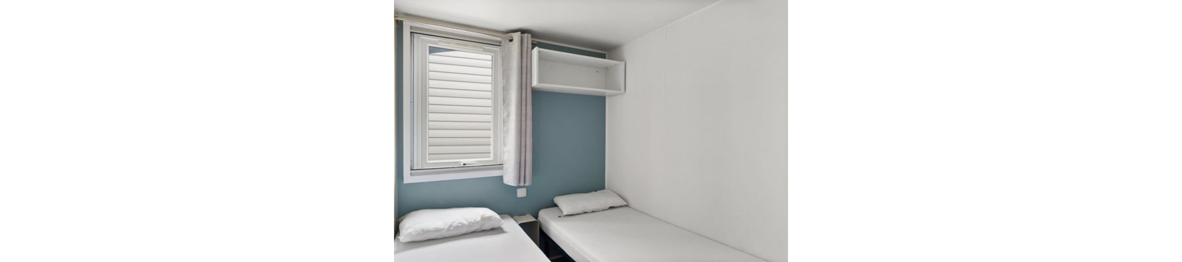 Compact bedroom interior featuring two simple single beds with white bedding. A window with light curtains lets in natural light, brightening the small space. Walls are painted white with a blue accent wall behind one bed, and a small open shelf is mounted above. The overall design is minimal and functional, suggesting a budget hotel room, hostel, or vacation rental.