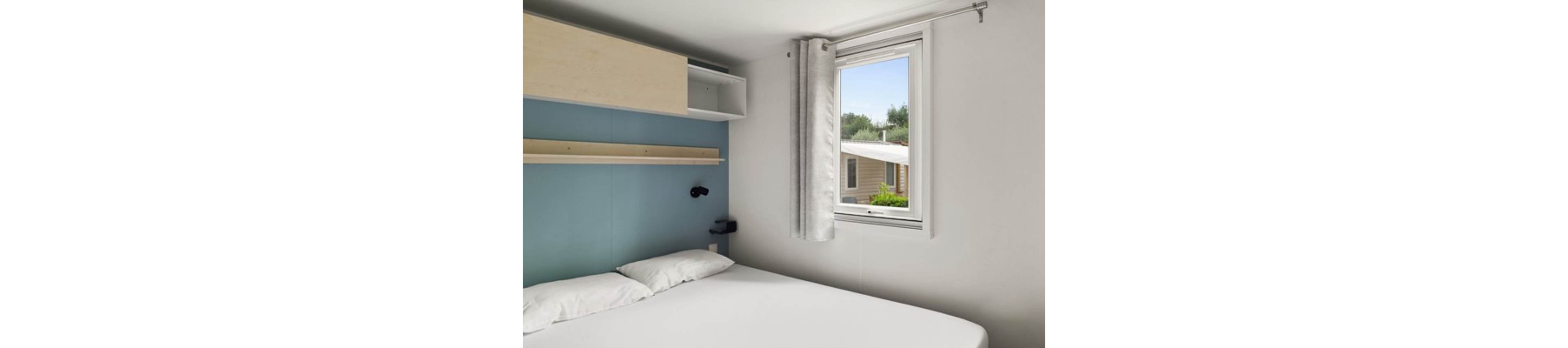 A small minimalist bedroom features a neatly made double bed with white linens. Above the bed, light wood shelves and cabinets provide simple storage against a pale blue accent wall. A window with light curtains lets in natural daylight and offers a view of a neighboring cabin and greenery outside. The overall scene feels clean, bright, and functional, suitable for a vacation rental or compact home.