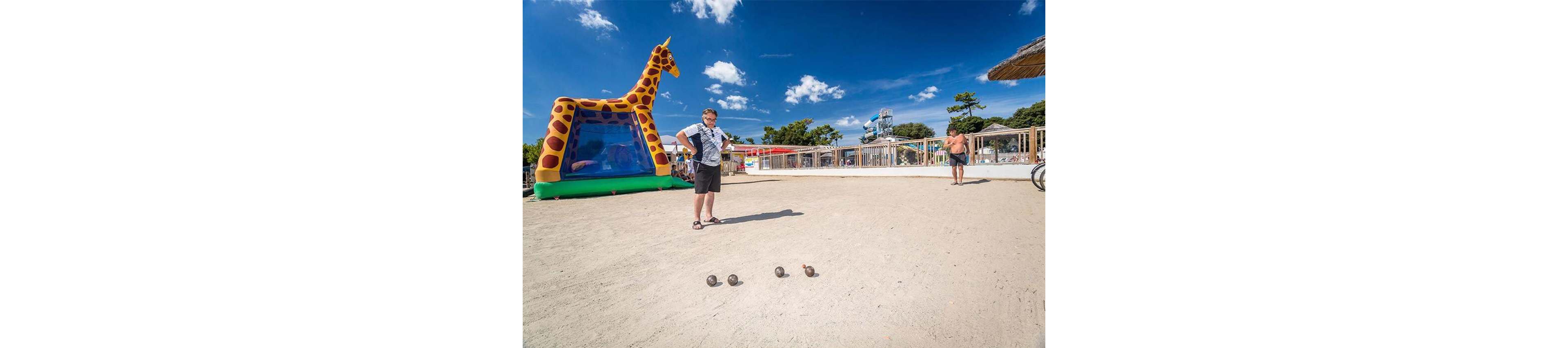 A man stands on a sandy court preparing to play pétanque, with several metal balls lined up in front of him. The scene is set in a sunny outdoor recreation area near the beach, featuring a large giraffe-shaped inflatable structure. Another person in swimwear walks in the background, and a bicycle is partially visible on the right. Bright blue sky with scattered clouds and distant water slides create a relaxed holiday atmosphere.