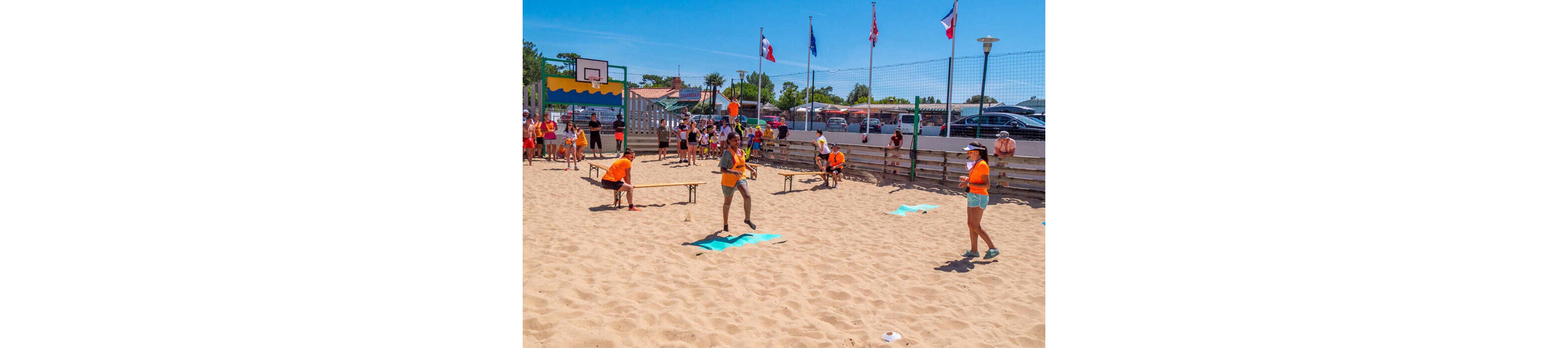 A group of people play a casual baseball-style game on a sandy beach court. Players in orange shirts are positioned around bases while one person prepares to hit the ball. Spectators watch from behind a wooden barrier and along the sides of the court. Flags fly above the fence and a basketball hoop is visible in the background under a clear blue sky.