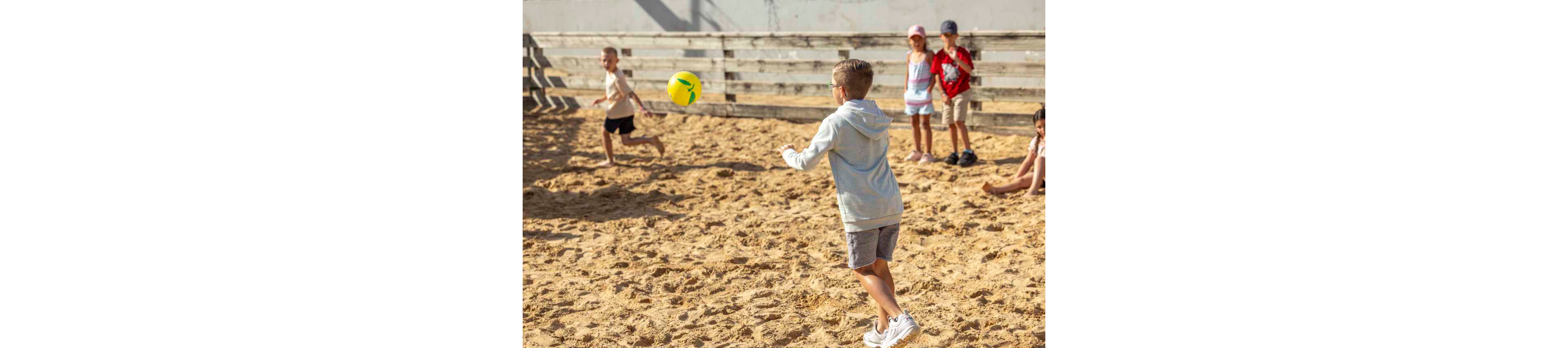 A group of children play with a yellow ball on a sandy outdoor court. One child in the foreground prepares to catch or throw the ball while others run and watch in the background. The scene is bright and sunny, suggesting warm weather and active playtime. A wooden fence surrounds the play area, giving the space a casual, playground-like atmosphere.