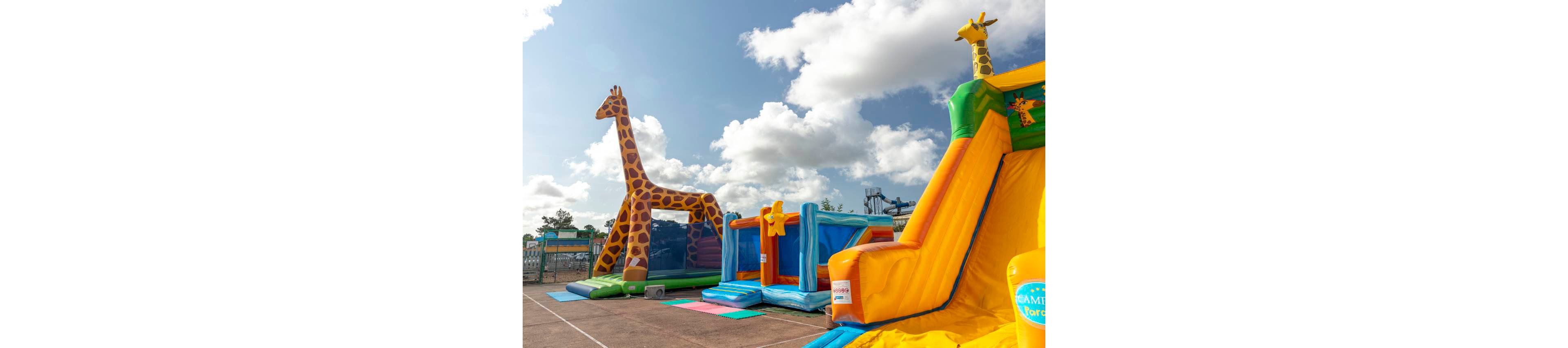 A bright inflatable playground features large giraffe-themed structures under a partly cloudy sky. Multiple bounce houses and slides in yellow, blue, and orange are set up on an open paved area. The scene suggests a funfair or amusement park environment with water slides visible in the background. The overall atmosphere is playful and family-friendly, ideal for children’s outdoor activities.