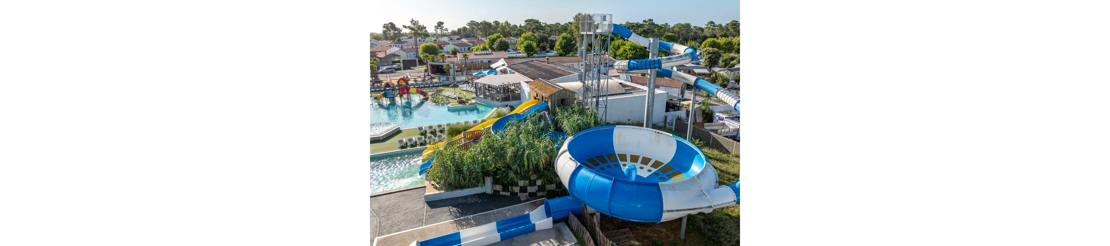 An outdoor water park features several large blue and white spiral water slides as its main attraction. The slides twist above a pool complex with sun loungers and shallow swimming areas. Greenery and tall grasses surround parts of the structure, softening the concrete and metal elements. In the background, low residential-style buildings and trees extend toward the horizon under a clear sky.