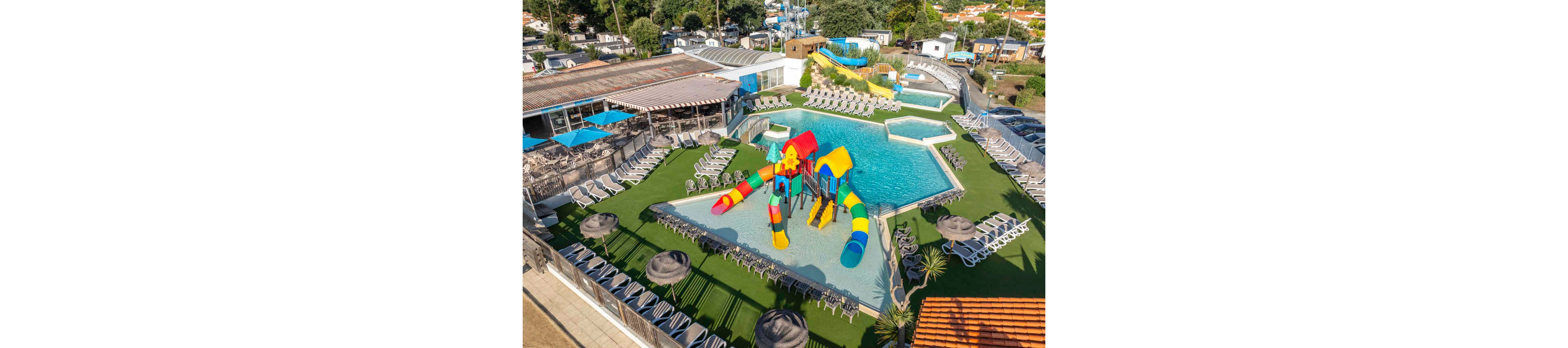 Aerial view of an outdoor resort pool featuring a colorful water playground with multiple slides. The pool is surrounded by rows of empty lounge chairs on artificial grass and sun umbrellas. Additional pools and water slides are visible in the background, along with nearby buildings and trees. The bright colors and clear weather create a fun, family-friendly vacation atmosphere.