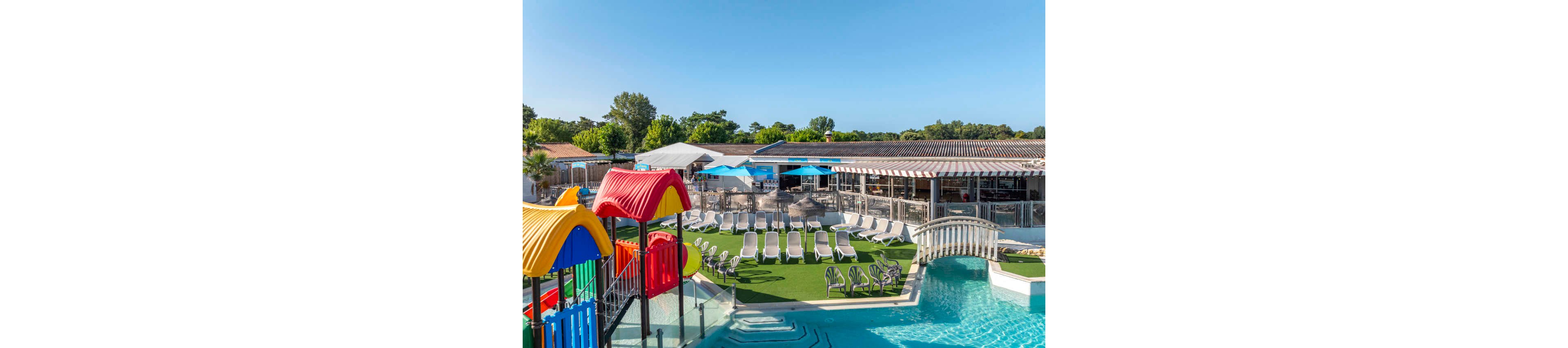 An outdoor resort pool area is shown under a clear blue sky. Numerous white sun loungers are arranged in rows on artificial grass beside the water. A colorful children's play structure with slides stands next to the pool, along with a small decorative bridge crossing the water. In the background, low buildings with terraces and umbrellas suggest a bar or restaurant area surrounded by greenery. The scene evokes a relaxed, family-friendly vacation atmosphere.