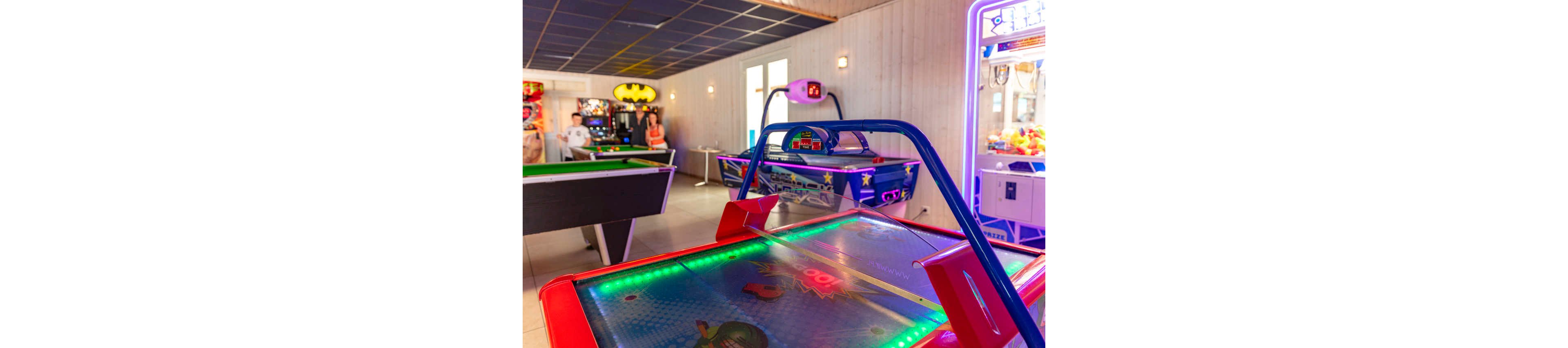 A brightly lit arcade room features a glowing air hockey table in the foreground. In the background, people play at pool tables and other arcade machines. Neon lights, colorful graphics, and illuminated game cabinets create a lively entertainment atmosphere. The setting appears casual and fun, suitable for leisure and socializing indoors.