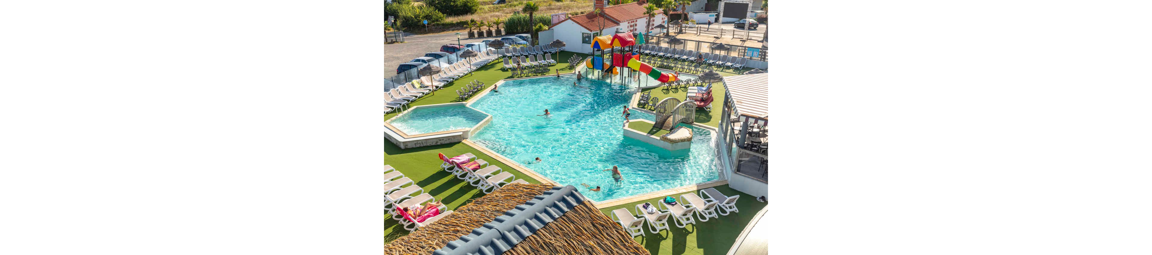 A large outdoor swimming pool complex features several interconnected pools surrounded by numerous sun loungers. Children and adults enjoy the water, with some playing near colorful slides and a small water playground structure. A separate shallow pool area is visible on the side, suggesting a kid-friendly zone. The scene is bright and sunny, creating a relaxed vacation atmosphere at a resort or holiday park.
