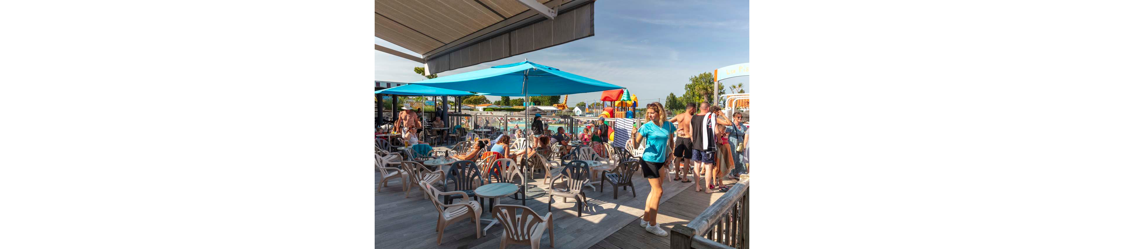 An outdoor café terrace is filled with people relaxing on plastic chairs under a large blue umbrella. The setting overlooks a busy swimming pool area with families and children enjoying the water. Colorful play structures and slides are visible in the background, adding to the lively atmosphere. The bright sunlight and clear sky create a warm, summery vacation vibe.