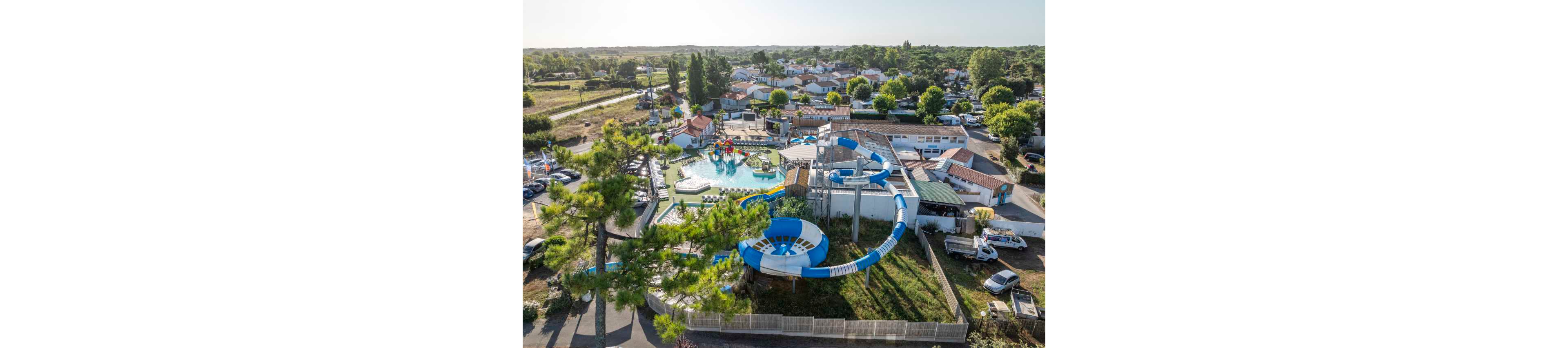 A large outdoor water park is captured from an aerial perspective within a residential neighborhood. The complex features twisting blue and white water slides, a splash play area, and a central swimming pool. Surrounding the park are parked cars, trees, and low-rise houses stretching into the distance. Bright sunlight and clear skies create a summery, family-friendly atmosphere.