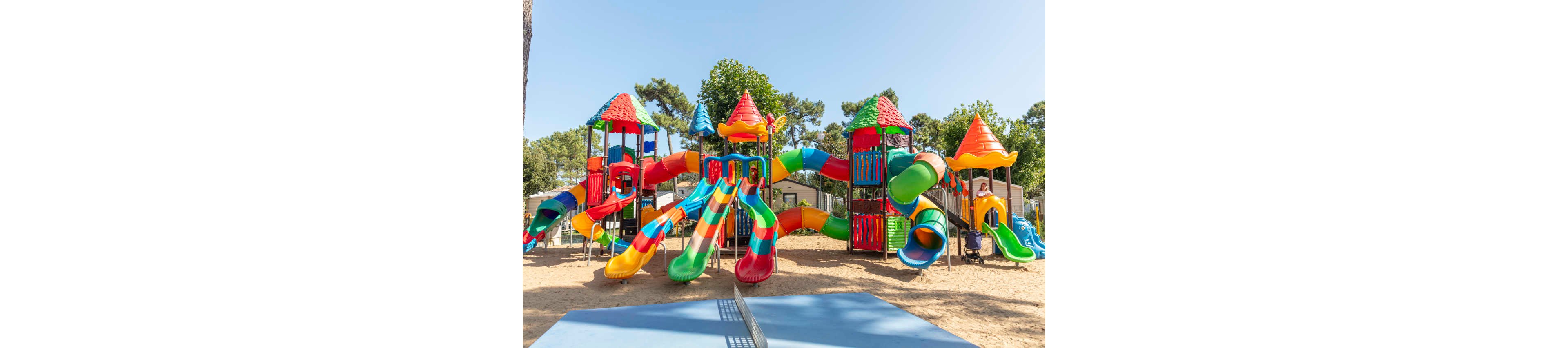 A large, colorful playground structure with multiple slides and towers stands in a sunny outdoor park. The equipment features bright red, green, yellow, blue, and orange elements, including twisting tube slides and open chutes. Trees and small buildings are visible in the background, suggesting a family-friendly recreation area or campground. The sandy ground and clear sky create a cheerful, inviting atmosphere for children to play.