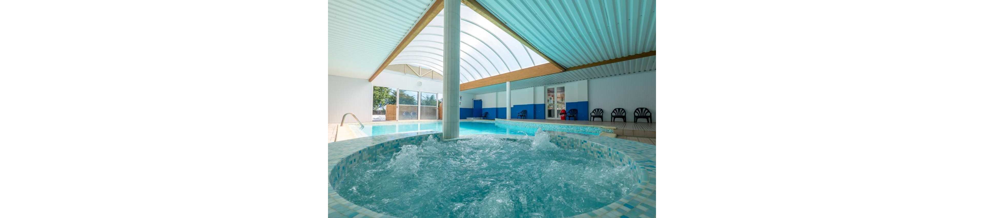 An indoor spa area features a bubbling circular jacuzzi in the foreground. Behind it, a rectangular swimming pool extends under a translucent arched roof that lets in natural light. White and blue walls, plastic chairs, and structural columns frame the space, creating a clean and modern look. The scene suggests a relaxing wellness or leisure environment in a resort or recreational facility.