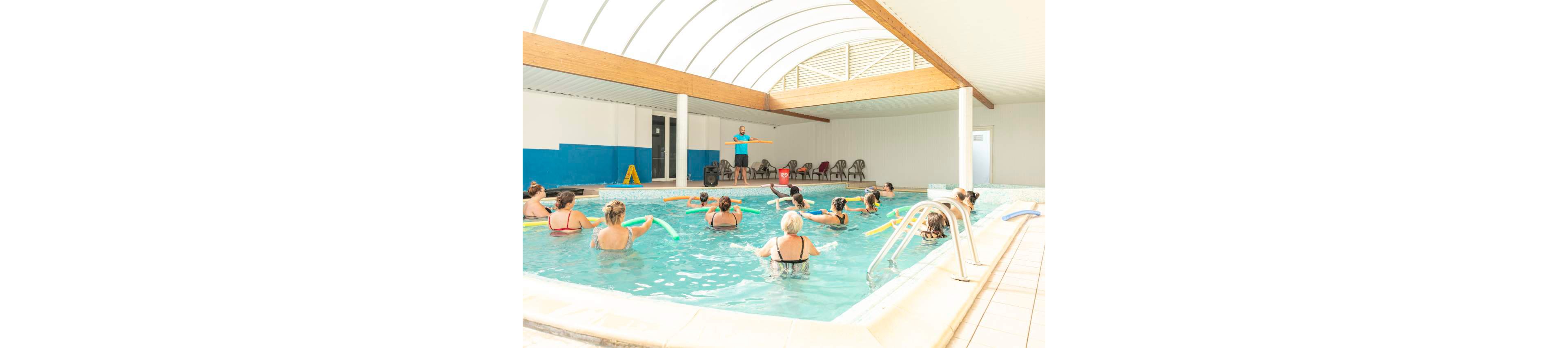 A group of adults participates in a water aerobics class in a bright indoor swimming pool. An instructor stands at the pool edge demonstrating exercises while the class follows along with foam pool noodles. The space features a curved translucent roof, white walls, and a row of empty chairs in the background. The atmosphere is active, social, and focused on low-impact fitness and wellness.