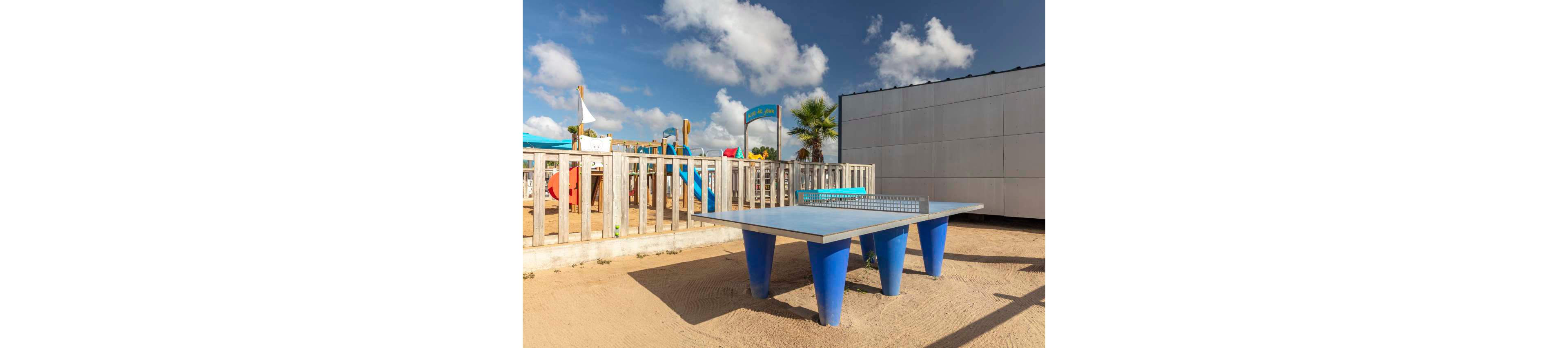 A blue outdoor ping pong table stands on a sandy surface in a recreational area. Behind it, a fenced playground with colorful equipment is visible under a bright sky with scattered clouds. A palm tree and a white modern building frame the background, suggesting a coastal or resort setting. The scene is sunlit and relaxed, emphasizing leisure and outdoor play.